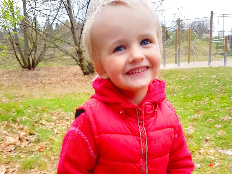 Toddler boy in a red jacket smiling outdoors near the TIMS playground